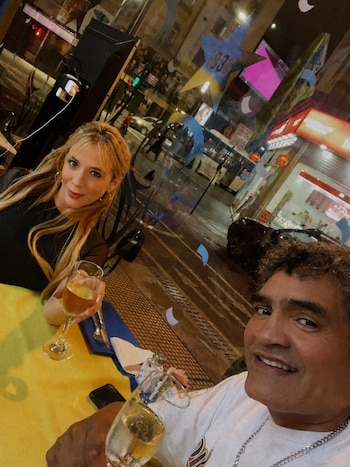 Un hombre de cabello oscuro y una mujer rubia sonríen en una mesa con copas de bebida frente a un ventanal hacia una calle nocturna