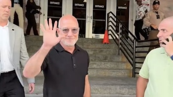 Jeff Bezos sonríe y levanta la mano derecha frente a las escaleras y puertas de vidrio de un edificio