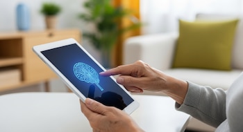 Manos de una persona mayor asiática con piel arrugada sostienen una tableta digital. En su pantalla oscura, un icono azul brillante de cerebro con circuitos.