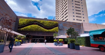 Fachada de un centro comercial con jardín vertical, letrero identificativo, peatones, maceteros y un edificio alto en el fondo
