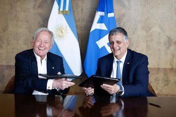 Dos hombres sonríen sosteniendo carpetas negras sobre una mesa. Detrás, se observan una bandera argentina y una bandera azul con el logo de YPF