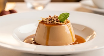 Primer plano de un postre cremoso color caramelo en un plato blanco hondo, cubierto con salsa de cajeta, nueces picadas y una hoja de menta.