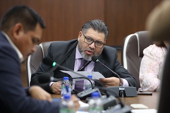 Hombre con barba y gafas, vestido de traje, habla en un micrófono de mesa mientras sostiene documentos. Se observan botellas de agua y otros participantes