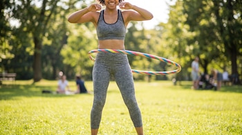 Mujer con ropa deportiva gris usa un hula hula multicolor a la altura de la cintura en un parque verde.