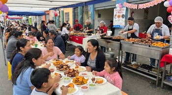 Una escena animada de varias familias con madres e hijos comiendo chicharrones peruanos en mesas blancas al aire libre, con puestos de comida y globos al fondo.