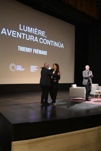 Thierry Frémaux y Gabriela Ricardes se abrazan en un escenario. Detrás, una pantalla muestra "LUMIÈRE, AVENTURA CONTINÚA". Otro hombre aplaude a la derecha