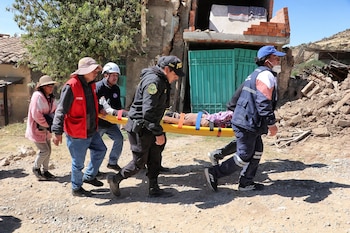 Seis personas, cuatro de ellas con chalecos y uniformes de rescate, cargan una camilla con un herido en un área con escombros y viviendas dañadas