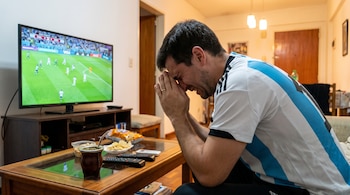 Hombre con camiseta de fútbol de Argentina se cubre la cara frente a un televisor que muestra un partido; un mate, patatas fritas y mandos a distancia están en la mesa.
