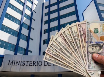 Múltiples pilas de monedas doradas apiladas y dispersas sobre una superficie de madera, con la bandera de El Salvador, que es azul y blanca, ondeando de fondo