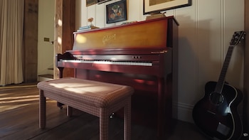 Un piano vertical rojo con la marca Gibson y un banco de tela a cuadros en un salón con suelo de madera. Una guitarra acústica negra se apoya en la pared adyacente