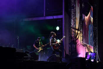 Músicos de rock en el escenario de un concierto, tocando guitarras eléctricas bajo luces verdes y púrpuras. Hay estructuras de truss, pantallas y altavoces visibles