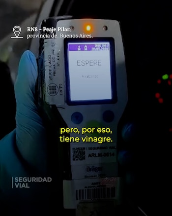 Una mano enguantada de azul sostiene un alcoholímetro profesional. La pantalla del dispositivo muestra la palabra "ESPERE" y un indicador luminoso naranja se enciende en la parte superior
