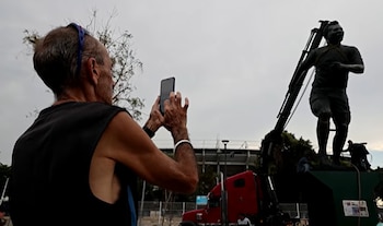 Un hombre de espaldas fotografía con su smartphone una estatua de atleta movida por una grúa, bajo un cielo nublado, con un estadio al fondo