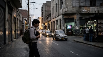 Un joven con mochila y cuaderno cruza una calle por la noche frente a varios taxis antiguos, persianas metálicas y un puesto de periódicos.