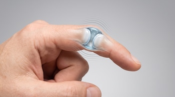 Una mano humana con un dedo flexionado muestra una articulación translúcida, huesos, líquido sinovial y burbujas de gas, con líneas que indican sonido.