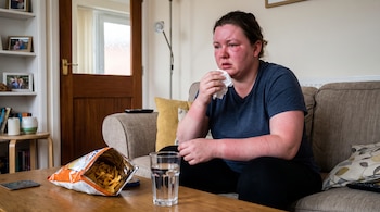Mujer sentada en un sofá con la cara y los brazos enrojecidos, sostiene un pañuelo de papel; en la mesa hay una bolsa de patatas fritas y un vaso de agua.