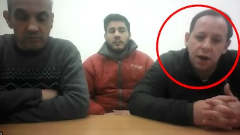 Tres hombres sentados en una mesa de madera, uno hablando, los otros dos observando, frente a una pared blanca