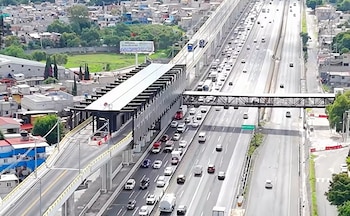 Vista aérea de una estación elevada y un puente peatonal sobre una autopista con tránsito de automóviles junto a una zona urbana