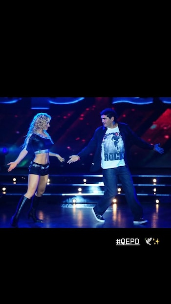 Un hombre y una mujer bailan en un escenario iluminado con luces azules y rojas, con el texto QEPD en la esquina inferior derecha