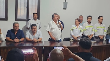 Un grupo de hombres y una mujer en una reunión. Un hombre mayor habla por micrófono. Algunos visten camisas con rayas reflectantes, otros lisas