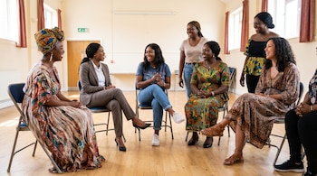 Siete mujeres afrodescendientes sentadas en sillas formando un círculo en una sala luminosa. Todas sonríen y están vestidas con diversos atuendos, algunos de ellos coloridos y estampados.