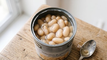 Lata de metal abierta con porotos blancos en líquido, colocada sobre una tabla de madera al lado de una cuchara