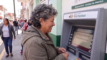Una mujer de cabello canoso frente a un cajero automático del Banco del Bienestar con la frase Pensión Mujeres Bienestar en pantalla.