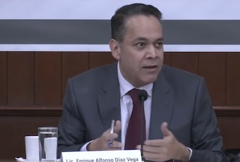 Enrique Díaz Vega, un hombre con traje gris y corbata roja, habla frente a un micrófono en una sala con paneles de madera, con un vaso de agua