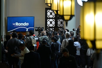 Un grupo de periodistas y camarógrafos capturan imágenes de una pantalla que muestra "La Ronda Conferencia de Prensa Presidencial", con lámparas decorativas borrosas