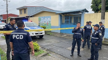 Agentes de la PCD en chalecos oscuros, un patrullero policial con luces encendidas, cinta amarilla "escena de crimen" y una escuela al fondo en San José.
