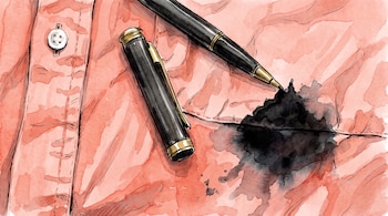 Una pluma negra con detalles dorados y su tapa junto a una mancha de tinta negra sobre una camisa rosa con un botón.