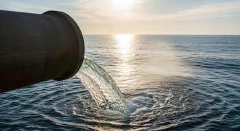 Agua transparente fluyendo desde una tubería de gran diámetro hacia la superficie del mar, con el sol reflejado en el agua a lo lejos.