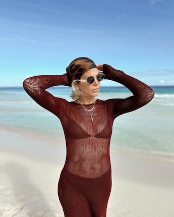 Jenny Williams posa en la playa con un vestido calado café, gafas de sol y un pañuelo en la cabeza frente al mar