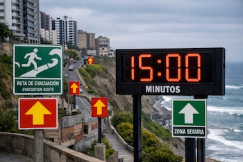 Reloj digital con quince minutos, señal de ruta de evacuación, señal de zona segura y múltiples flechas de dirección. Carretera costera, edificios y mar.