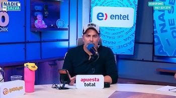 Un hombre con gorra y camiseta negra habla ante un micrófono sentado a una mesa de estudio de transmisión con pantallas de fondo