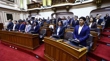 Sala de sesiones con legisladores de pie frente a pupitres de madera equipados con micrófonos y pantallas. Alfombra roja cubre el suelo