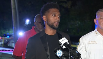 Brandon Scott habla ante varios micrófonos junto al comisario R. Worley de noche, con luces de patrulla al fondo