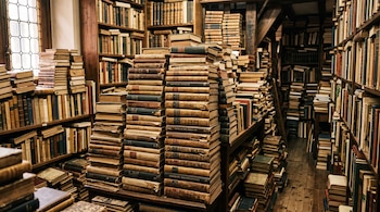 Pilas de libros antiguos sobre mesas de madera y estanterías repletas de tomos encuadernados.