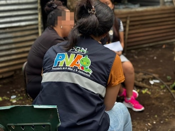 Vista trasera de una persona con chaleco azul de la PNA y logo de un sapo verde, conversando con otra persona sentada en un entorno informal