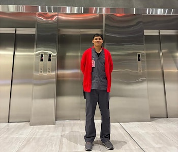 Hombre joven con cárdigan rojo y uniforme gris oscuro de pie. Detrás de él, tres ascensores de acero inoxidable con indicadores de piso y botones