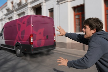 Un hombre con sudadera gris extiende los brazos hacia una furgoneta morada en movimiento frente a un edificio de fachada blanca.