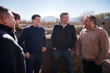 Cuatro hombres sonríen y conversan al aire libre. Hay fardos de heno, un edificio y montañas en el fondo. El cielo está despejado