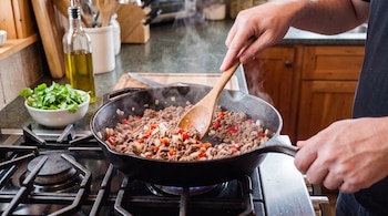Manos revolviendo carne picada con cebolla y morrón en una sartén sobre la hornalla, con una espátula de madera, cerca de hierbas frescas y aceite.