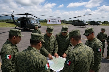 Soldados con uniforme de camuflaje y parches de México observan un mapa sobre una pista junto a varios helicópteros militares.