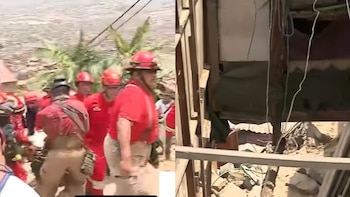 Bomberos con cascos y uniformes rojos en una ladera, junto a una estructura precaria de madera con cables colgantes y escombros