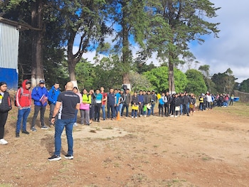 Un grupo de personas, mayormente jóvenes, se encuentra en fila al aire libre escuchando a un instructor. Hay árboles y estructuras en el fondo bajo un cielo nublado