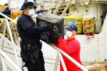 Dos hombres con uniformes, mascarillas y guantes manipulan un fardo rectangular oscuro en la cubierta de un barco. Uno lleva una gorra de la Guardia Costera