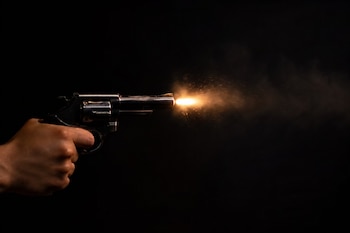 Mano empuñando un revólver plateado del cual sale una llamarada y chispas sobre un fondo negro.