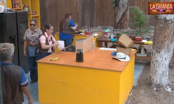 Varias personas en una cocina rústica al aire libre con encimeras amarillas y paredes de madera. Una mujer sostiene un recipiente mientras otra camina detrás de un refrigerador