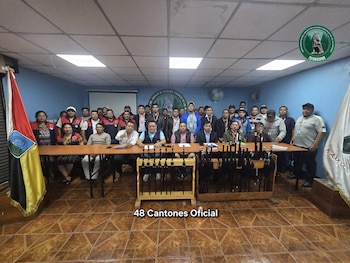 Un grupo de personas posa detrás de una mesa de madera en una sala con paredes azules, banderas en los laterales y bastones de mando al frente